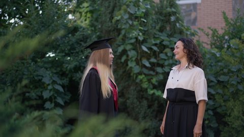 Excited grad woman throwing graduation cap up in slow motion with cheerful mother smiling clapping standing on backyard outdoors. Portrait of happy Caucasian parent and daughter celebrating
