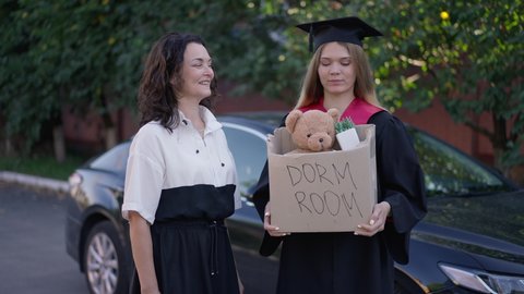 Young graduate woman and mature mother talking smiling standing at car with dorm room box. Portrait of Caucasian daughter and parent outdoors on graduation day. Slow motion
