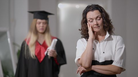 Portrait of stressed Caucasian sad mother standing at home with blurred graduate daughter at background. Upset woman thinking as cheerful millennial lady rejoicing with rolled diploma