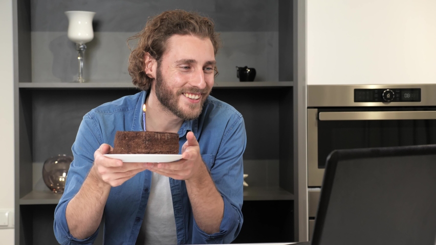 Happy man celebrating birthday alone. Handsome male bday with party cake and blowing candle. Guy using laptop webcam having videocall with friends, nice mood, positive emotion blows out the candle.