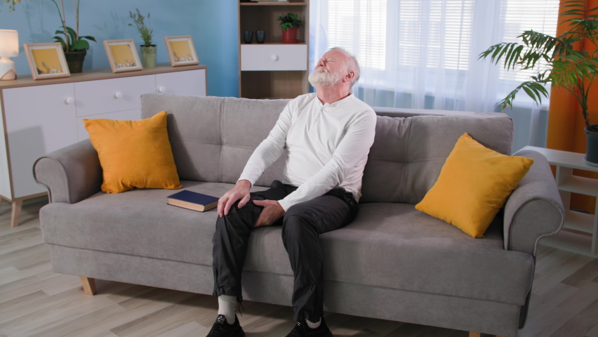 Old man reads book then gets up from the couch and feels pain in his leg, granddad with osteoporosis rubs his knee at home | Shutterstock HD Video #1083607054