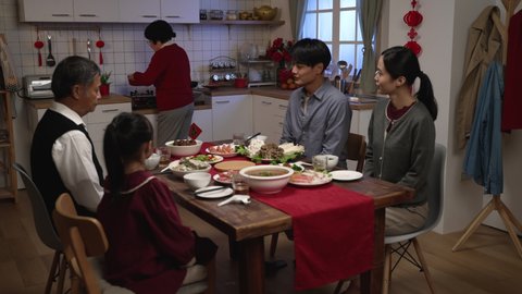 grandmother wearing red clothes serving delicious food on dining table as her family members clapping hands for her. getting ready for big meal on chinese lunar new year's eve. translation: spring