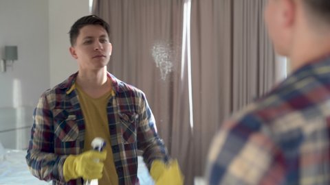 House cleaning. Caucasian man in casual clothes cleans the mirror from dust and dirt using spray and a yellow napkin. Rubber gloves on hands.