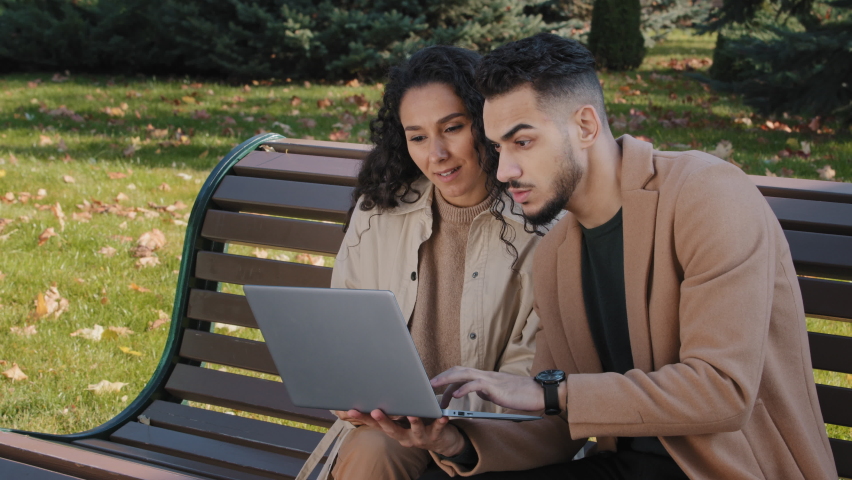 Excited hispanic couple reading good news on laptop sitting on bench in autumn park successful partners happy hugging surprised to receive email man winner celebrating victory lottery embracing woman