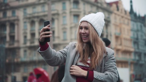 Portrait of young woman during facetime call on the street. Girl sending best wishes to family and friends on christmas standing in front of beautiful european architecture. Concept of travel, gadgets