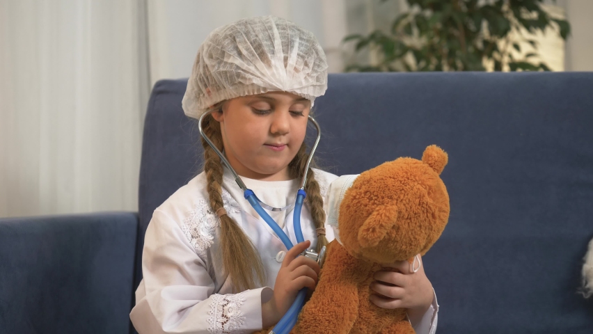 Little girl in white doctor's coat is playing. child plays doctor. girl using stethoscope treating teddy bear sitting in an isolated room. small child dreams of becoming doctor by helping teddy bear