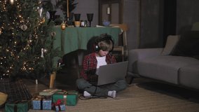 Little kid boy using laptop computer sitting near christmas tree with headphones, playing computer games, listening to music, doing shopping online, making payment on christmas. Child boy with laptop