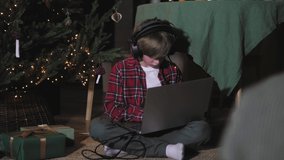 Positive energetic child boy with headphone listening to music on laptop or computer on christmas, excited about christmas playlist, sitting under christmas tree at night