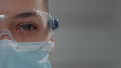 Scientific researcher looking at camera with glasses and face mask for protection. Biochemist wearing protective goggles and showing half of face in microbiology laboratory. Close up