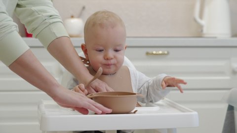 Toddler learning to eat with a spoon, smiling kid with food stained face, infant baby sits on feeding high chair, stained with porridge. High quality 4k footage