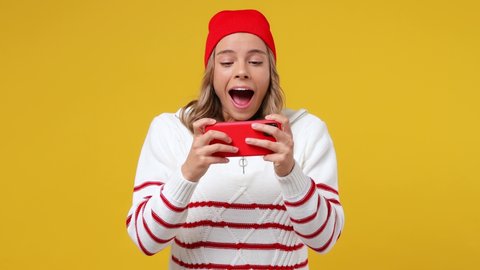 Gambling fun young girl teen student wears striped white shirt hat using play racing on mobile cell phone hold gadget smartphone for pc video games isolated on plain yellow background studio portrait