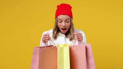 Beautiful excited happy vivid young girl teen student wears striped white shirt hat holding looking into package bags with purchases after shopping isolated on plain yellow background studio portrait