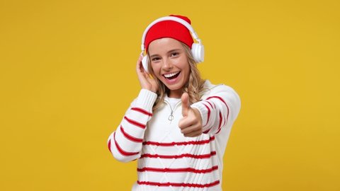 Beautiful cheery happy blithesome young girl teen student wears striped white shirt hat listen music in headphones dance show thumb up like gesture isolated on plain yellow background studio portrait