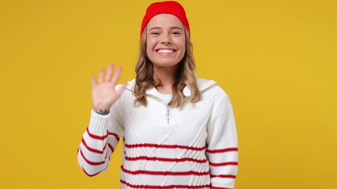 Beautiful cheery young girl teen student wears striped white shirt hat look around for friend find waving meet greet with hand as notices someone isolated on plain yellow background studio portrait