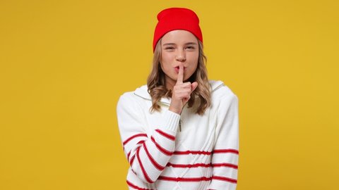 Secret young girl teen student wears striped white shirt hat look aside say hush be quiet with finger on lips shhh gesture isolated on plain yellow background studio portrait. People emotions concept