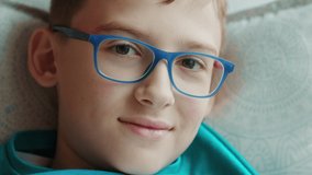Cute caucasian kid in modern home interior. Portrait of a cute schoolboy wearing blue eyeglasses and looking serious, while laying on a couch. High quality 4k footage