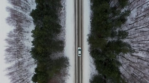 Aerial top down view from drone of white sedan vehicle driving on snowy ice asphalt road, landscapes in winter. Birds eye view of car moving on area surrounded by coniferous forest shelter-belt