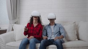 Excited Hispanic young couple wearing virtual reality headsets sitting on the couch playing a tennis game with the consoles on weekend at home