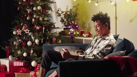 African American man speaking on video call on smartphone, sitting on sofa. Black afro guy speaking with friends online in app. Christmas time concept. New Year holidays. Cozy winter evening at home.