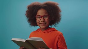 African American school girl in eye glasses reads book and smiles at camera standing on blue background. Smart kid enjoys reading paper books. - Powered by Shutterstock - Get 15% off with code: PIKWIZARD15