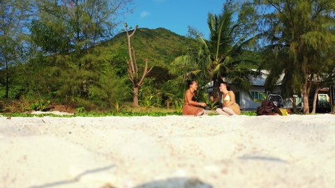 Two young women chatting on calm exotic beach with white sand under palm trees of tropical island in Ko Pha Ngan, Thailand