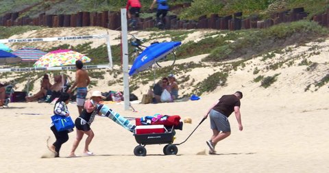LOS ANGELES, CALIFORNIA, USA - JULY 18, 2021: Young family moves their belongings in cart walking on beach sand near volleyball game in Los Angeles, California, 4K