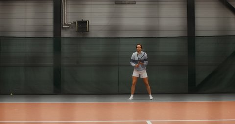 A young woman tennis player in a stylish sports uniform runs on the tennis court, deftly hitting a tennis ball with a racket. Video filming through a net stretched in the middle of a tennis court
