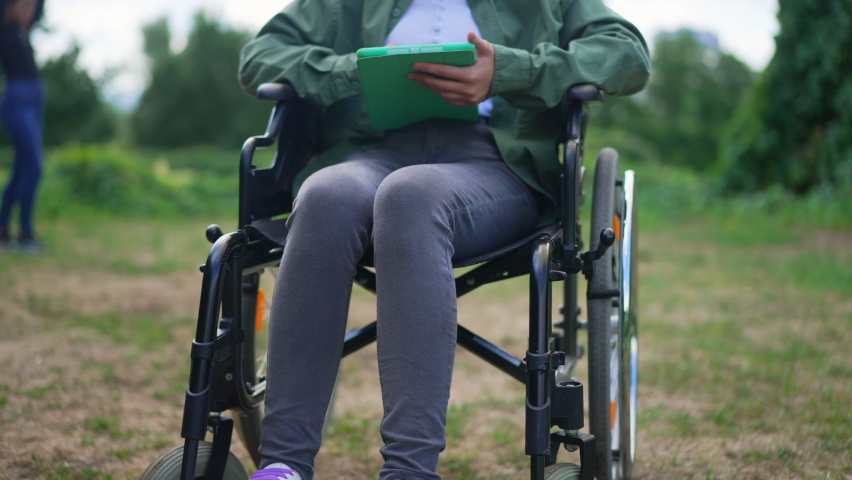 Unrecognizable girl in wheelchair with tablet resting in summer park after accident. Caucasian teenager surfing Internet in slow motion outdoors. Trauma and lifestyle concept.