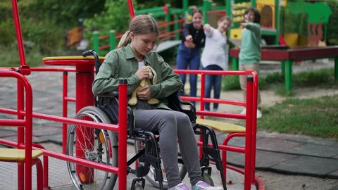 Crying desperate Caucasian girl in wheelchair hugging toy as blurred children pointing laughing at background. Portrait of stressed bullied child on playground with rude kids. Slow motion.