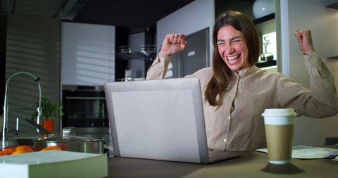Cinematic shot of young successful businesswoman is controlling emails on laptop while working in kitchen from home and exulting and celebrating a successful deal or promotion in her career.
