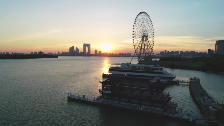 Aerial cityscape of a ferris wheel, boats and skyline in the sunset on Jingji lake, Suzhou, China. Camera moves forward