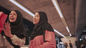 Two cheerful asian muslim girls look away showing their paper shopping bags while stand on pedestrian concrete bridge in nighttime, an online vlog video making, midnight sale, eco friendly concept