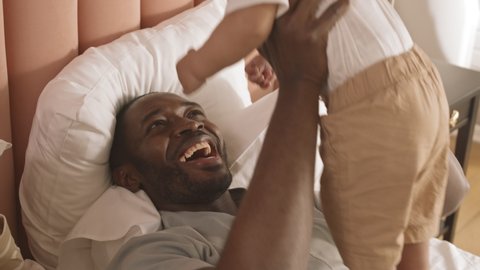 High-angle of excited young father lying in bed, holding little child, lifting his up and putting down, smiling