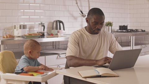 Waist-up of occupied African man working at kitchen table, typing on laptop computer, his Biracial baby sitting in high chair nearby