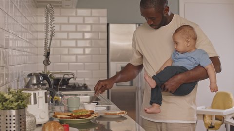 Medium shot of young single parent holding adorable toddler with one hand and making sandwich in kitchen