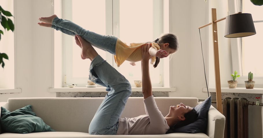 Overjoyed millennial mom lying on cozy couch on back holding little daughter in air on legs bent in knees play game imitate flight. Carefree child girl preschooler act flying doing gymnastics with mom Royalty-Free Stock Footage #1083679558