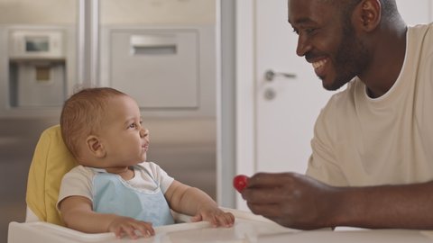 Medium close-up of happy African-American father sitting at table in kitchen, feeding his Biracial baby with fresh strawberry
