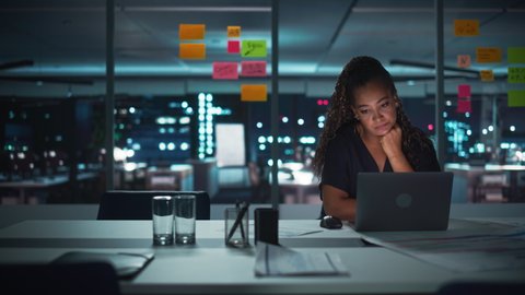 Overworked African American Businesswoman Working on Laptop Computer in Big City Office Late in the Evening. Tired Stressed Female Entrepreneur trying to Find Solution for Business Problems.