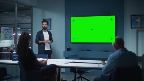 Company Operations Manager Holds Meeting Presentation for a Team of Economists. Adult Male Uses Wide TV Set with Horizontal Green Screen Mock Up Display. People Work in Business Office.