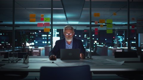 Successful Senior Businessman Working on Laptop Computer in Big City Office Late in the Evening. Investment Analyst Checking Financial Graphs from Project Management Reports.