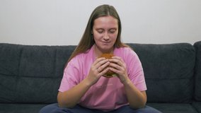 Plump young woman with blue eyes and pink T-shirt is sitting on gray sofa and eating big fat burger with great pleasure. Cholesterol, junk food, alcohol, unhealthiness, gluttony fat