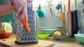 Woman grinds orange carrots on silver metal grater in the kitchen on blue board for cooking. In the background there are colorful plates with vegetables. Healthy food, vegetables, cooking at home
