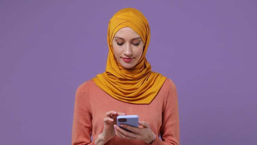 Calm young arabian asian muslim woman in abaya hijab yellow clothes hold use talk on mobile cell phone conducting pleasant conversation isolated on plain pastel light violet background studio portrait