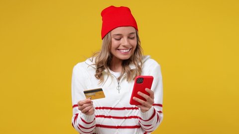 Young girl teen student wears striped shirt hat using mobile cell phone hold in hand credit bank card doing online shopping order delivery to home isolated on plain yellow background studio portrait