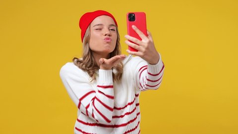Beautiful cheery happy vivid young girl teen student wears striped white shirt hat doing selfie shot on mobile phone post photo on social network isolated on plain yellow background studio portrait