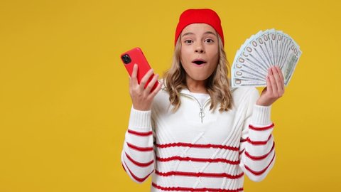 Excited joyful charming fun young girl teen student wears striped white shirt hat using mobile cell phone hold fan of cash money in dollar banknotes isolated on plain yellow background studio portrait