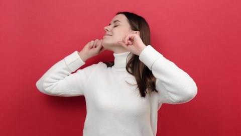 Portrait of serious young brunette woman 20s keeping ears closed not to listen loud annoying noise, high decibel sound, wears white sweater, posing isolated over red color background wall in studio