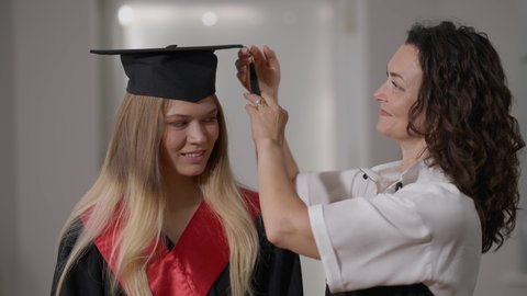 Smiling mother putting graduation cap tassel right to left on cap of happy daughter standing indoors. Proud Caucasian woman and graduate at home. Family support and education concept