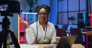 Portrait of lovely smiling confident high-skilled modern african businesswoman in stylish wear in glasses which sitting in front of camera in evening office and recording video