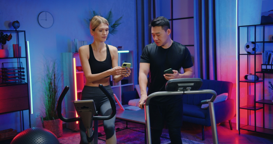 Active lifestyle concept where attractive smiling active sporty blond woman and asian man exercising at home together using running track and stationary bike and talking between themselves